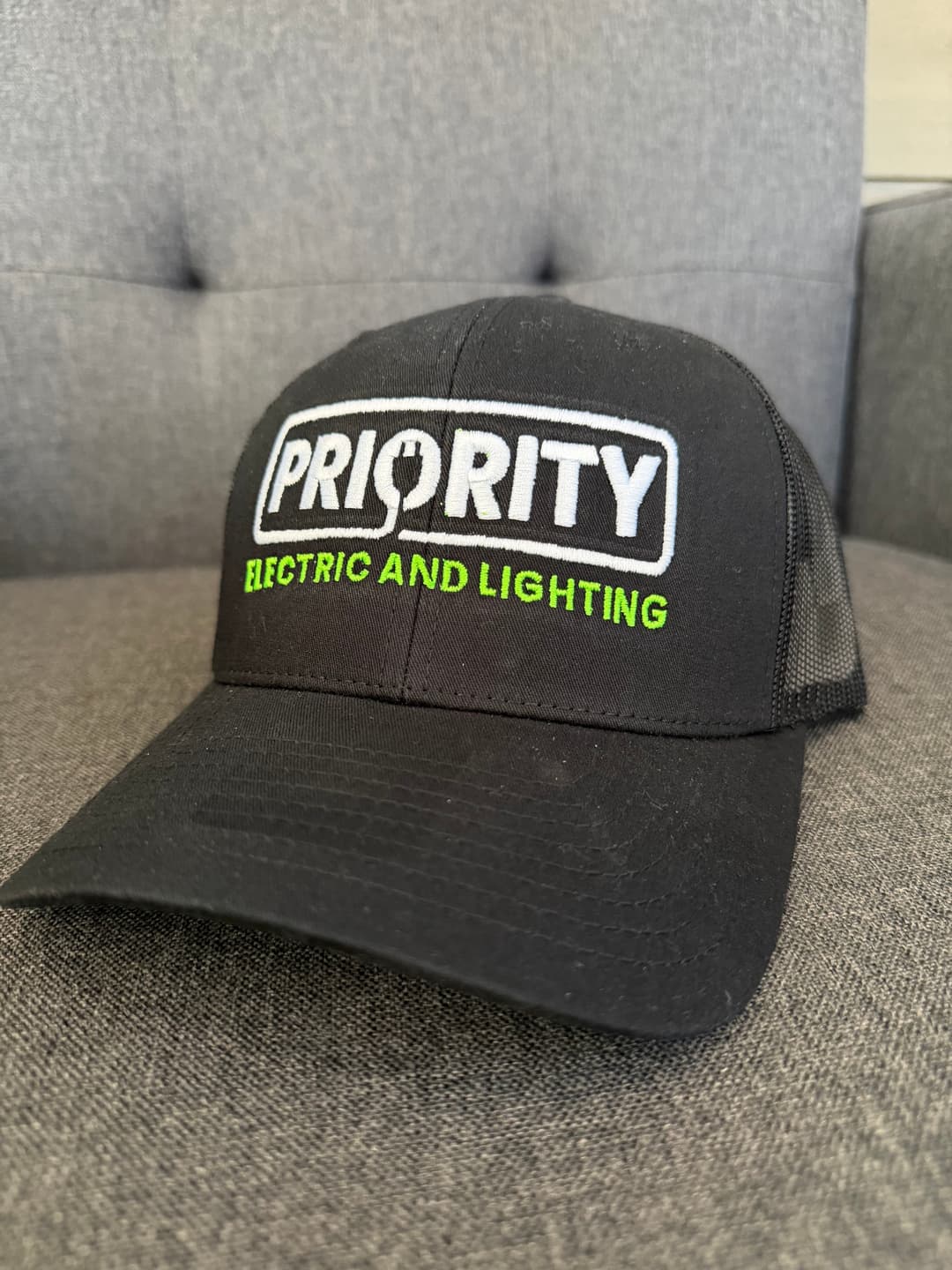 Black trucker hat with "Priority Electric and Lighting" logo in green and white embroidery.