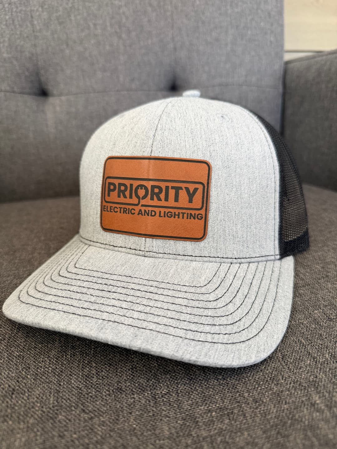 Gray snapback hat with brown leather patch for Priority Electric and Lighting on a gray background.